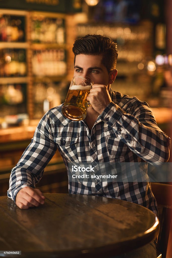 Young man drinking beer in a bar. Pensive young man sitting alone at the table in a pub and drinking beer. Alcohol - Drink Stock Photo