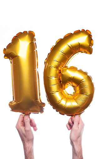Gold foil number 16 balloons held by a caucasian male hand. The numbers are being held at the base. The numbers are made from shiny golden foil and is inflated. The background is plain white.
