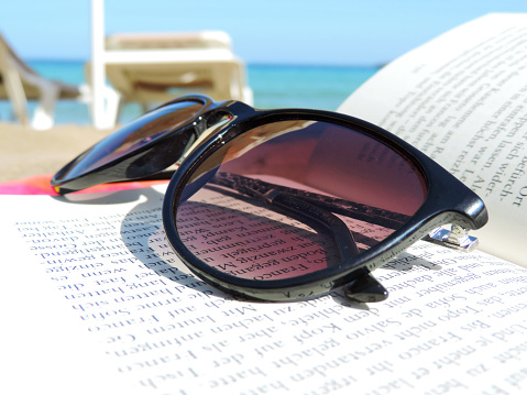 San Carlos, Spain - June 17, 2014: Sunglasses and book on the beach with selective focus and sun chairs in the background. Beach life scene with turquoise sea.
