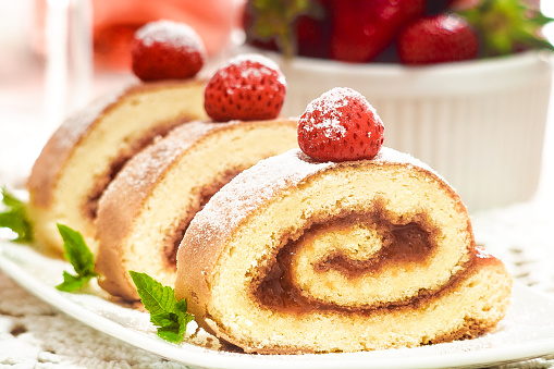 Slices of swiss roll with strawberry served on white plate daecorated with mint leaves, fresh strawberry and powder sugar.