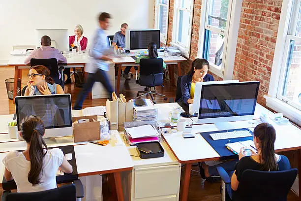 Wide Angle View Of Busy Design Office With Busy Workers At Desks Working.