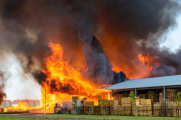 A factory in flames. Black smoke is billowing up from orange flames. Pallets of items are sitting outside the factory near the flames.