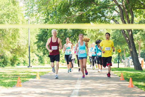 Diverse adults are excitedly running toward yellow finish line during marathon or 5k race for charity. They are running in group on footpath in park on sunny day. Senior and young adults are wearing athletic clothing and race contestant numbers.