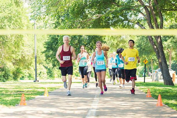 Diverse runners racing excitedly toward finish line during marathon Diverse adults are excitedly running toward yellow finish line during marathon or 5k race for charity. They are running in group on footpath in park on sunny day. Senior and young adults are wearing athletic clothing and race contestant numbers. walking for a cause stock pictures, royalty-free photos & images