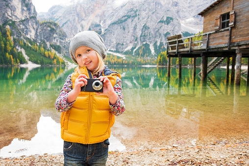 A young, blonde girl is standing on the shores of Lake Bries, smiling and tilting her head, holding a digital camera up. In the background, the water provides a mirror reflection of the autumn scenery, including autumn leaves, the Dolomites, and a rustic wooden pier with a small house on the pier.