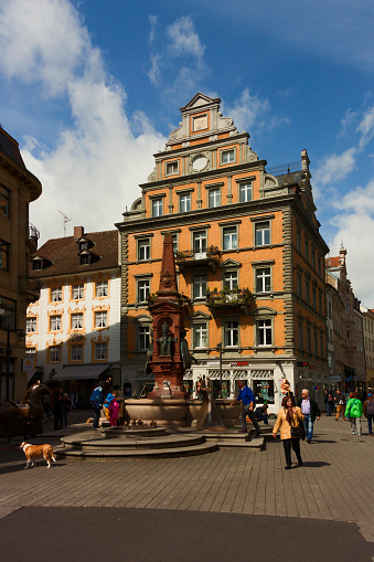 Constance, Germany - May 12, 2014: City view of Constance in germany. The tourist hub of Constance – Marktstätte square – extends towards the west. Built on land reclaimed from the lake, it was originally called Markt am Gestade (market on the waterfront). Haus Zum Korb, on the northern side of the square, is a late medieval house with Gothic windows. In the photo, many people can be seen walking in the shops in the town square. The imperial fountain (Kaiserbrunnen) was created by sculptor Hans Baur in 1897 but lost its original decorative fi gures in the 1940s. Professor Gernot Rumpf and his wife Barbara redesigned the monument and concealed within it a number of allusions to Constance’s past.