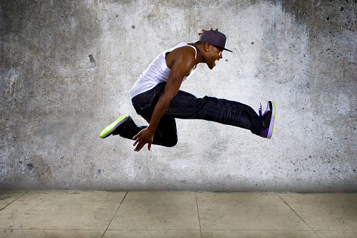 Black urban hip hop dancer jumping high on a concrete background.  The muscular man is athletic and active.  The concrete wall looks like an urban setting.  The man is break dancing or doing parkour.