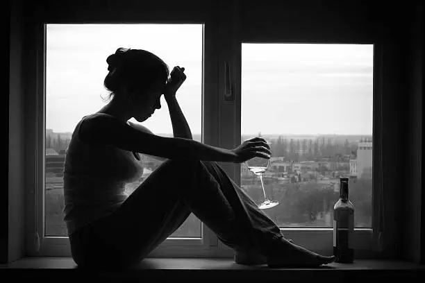 Sad young woman sitting on the window, drinks wine. Alcoholism problem. Black and white photography.