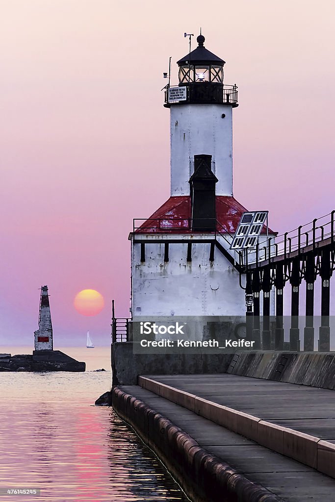 Idyllic Lighthouse Sunset The East Pierhead light of Michigan City, Indiana stands at the end of a long breakwater with elelvated metal catwalk approach as the sun sets on Lake Michigan with a silboat on the distant horizon and the smaller West Pierhead light across the channel opening. Lighthouse Stock Photo