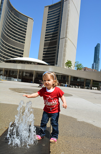 Beautiful Caucasian blond blue eyed 2 year old girl plays with a fountain by New City Hall at Nathan Phillips Square, Toronto, Ontario, Canada. 