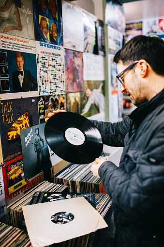 An mid adult, caucasian male customer examines a second hand vinyl record from stock on a shelf in a record store, vertical composition. 