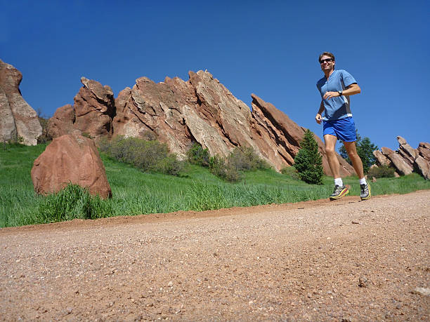 During hot and humind June morning, a man in shorts and sunglasses trail runs past the red rock Fountain Formation in Roxborough State Park Colorado.