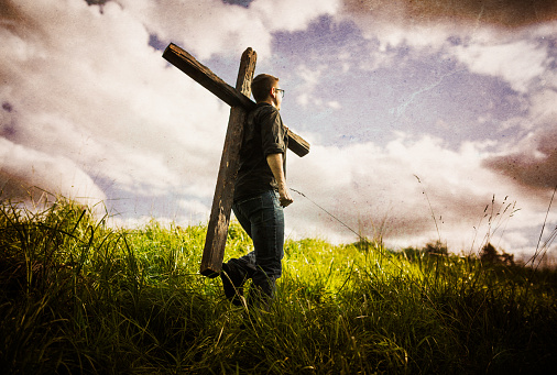 A man walks through a lush green wilderness area with blue sky and clouds overhead, carrying a large wooden cross over his shoulder.  A depiction of a Christian image given in the Bible, of picking up ones cross and following Jesus Christ.  Horizontal image with copy space in the grass.  An adventure or journey of discovery. Worn grunge paper texture overlaid on image for depth and character.