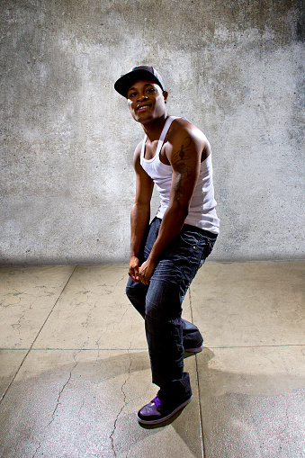 Photo of a muscular black man posing or dancing.  The man is on a concrete background that looks like an urban setting.  He is demonstrating dance moves and hip hop dance style choreography.
