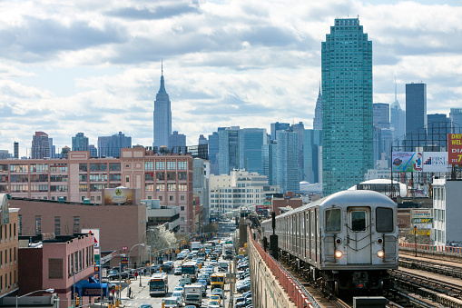 Train approaching  elevated subway station in Queens, New York. Finanancial buildings and New York skyline are seen in the background, on the left below can be seen a busy street full of cars at rush hour, cloudy, dramatic sky, horizontal orientation, USA