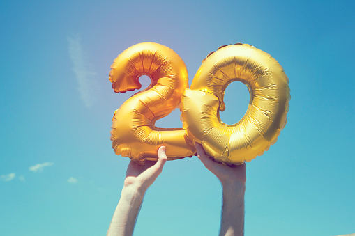 A gold foil number 20 balloon is held high in the air by caucasian male hand.  The image has been taken outdoors on a bright sunny day, the sky is blue with some clouds. A vintage style effects has been added to the image.