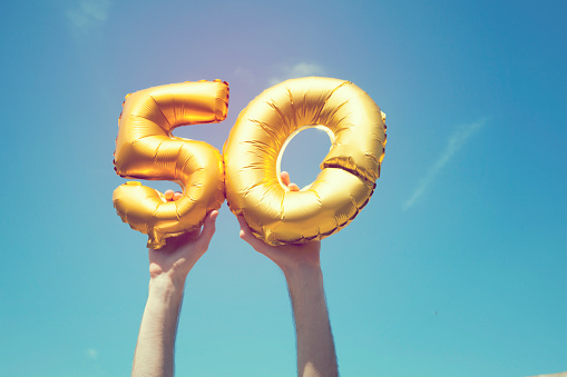 A gold foil number 50 balloon is held high in the air by caucasian male hand.  The image has been taken outdoors on a bright sunny day, the sky is blue with some clouds. A vintage style effects has been added to the image.