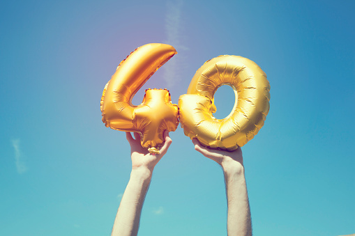 A gold foil number 40 balloon is held high in the air by caucasian male hand.  The image has been taken outdoors on a bright sunny day, the sky is blue with some clouds. A vintage style effects has been added to the image.