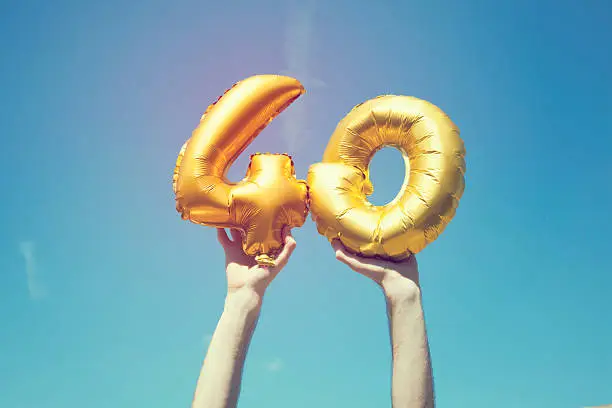 A gold foil number 40 balloon is held high in the air by caucasian male hand.  The image has been taken outdoors on a bright sunny day, the sky is blue with some clouds. A vintage style effects has been added to the image.