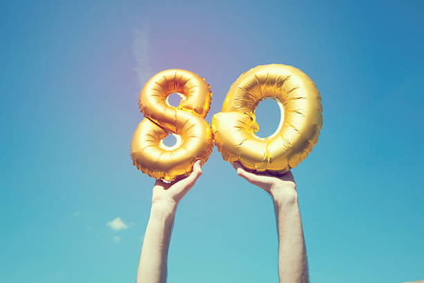 Gold number 80 balloon A gold foil number 80 balloon is held high in the air by caucasian male hand. The image has been taken outdoors on a bright sunny day, the sky is blue with some clouds. A vintage style effects has been added to the image. senior birthday card stock pictures, royalty-free photos & images