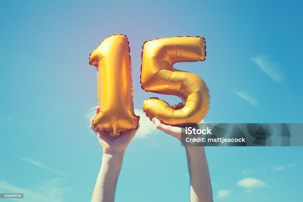 Gold number 15 balloon A gold foil number 15 balloon is held high in the air by caucasian male hand.  The image has been taken outdoors on a bright sunny day, the sky is blue with some clouds. A vintage style effects has been added to the image. Number 15 Stock Photo