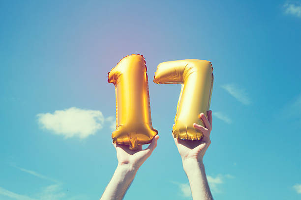 Gold number 17 balloon A gold foil number 17 balloon is held high in the air by caucasian male hand.  The image has been taken outdoors on a bright sunny day, the sky is blue with some clouds. A vintage style effects has been added to the image. number 17 stock pictures, royalty-free photos & images