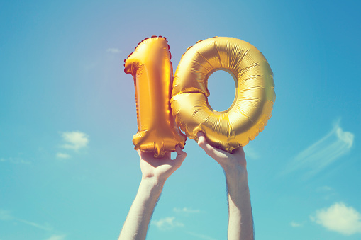 A gold foil number 10 balloon is held high in the air by caucasian male hand.  The image has been taken outdoors on a bright sunny day, the sky is blue with some clouds. A vintage style effects has been added to the image.