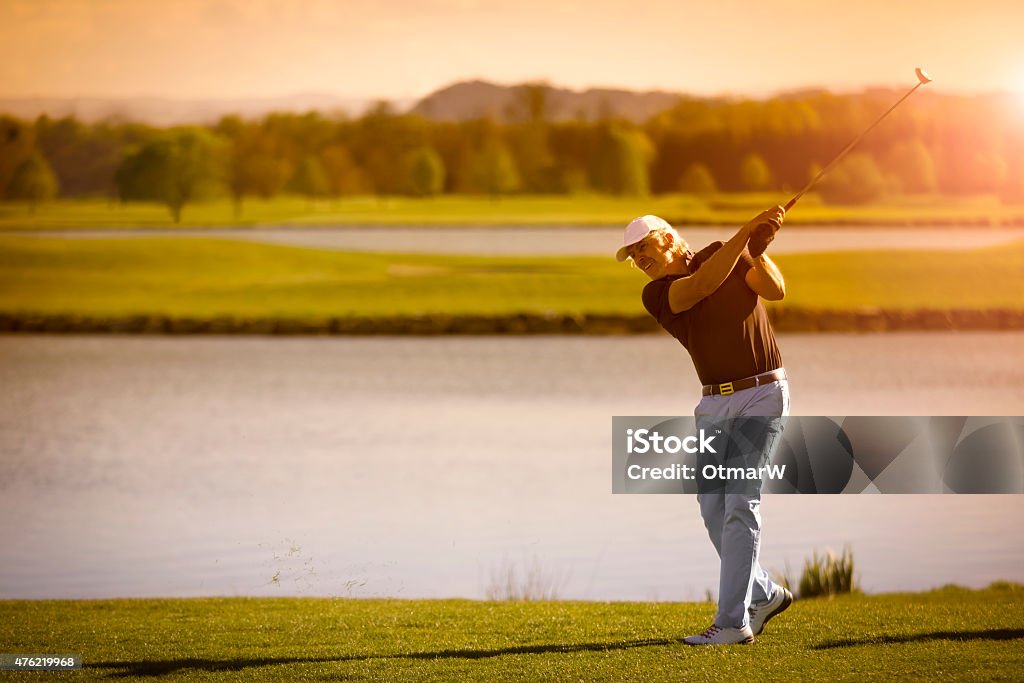 Senior golf player with copyspace. Male senior golf player swinging golf club with lake in background at sunset with copyspace. Golf Stock Photo