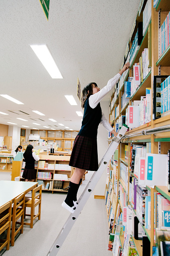 A view of a Japanese school library, book filled shelves. Interior shot, one female student has climbed a ladder to reach a book on an upper shelf. Two students are visible in the background. Interior, vertical composition. 