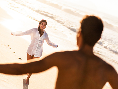 Happy Hispanic woman with her arms outstretched running towards her boyfriend. They are enjoying in summer day at the beach.