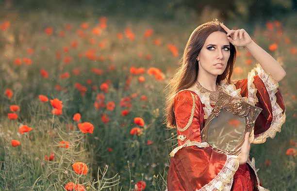 Beautiful Princess Holding Mirror in Summer Floral Landscape Portrait of a happy beautiful queen in red royal dress in a flower field mirror women baroque style fashion stock pictures, royalty-free photos & images