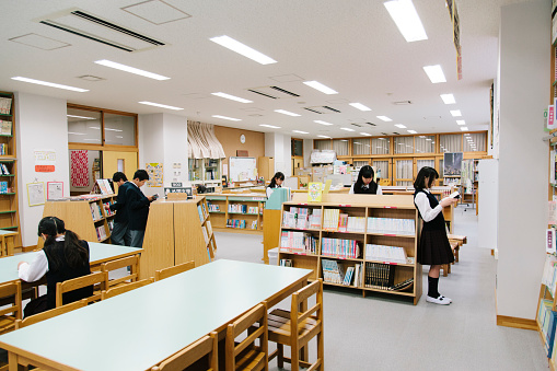 A view of a Japanese school library, book filled shelves. Interior shot, six students use the school library. Interior, horizontal composition. 