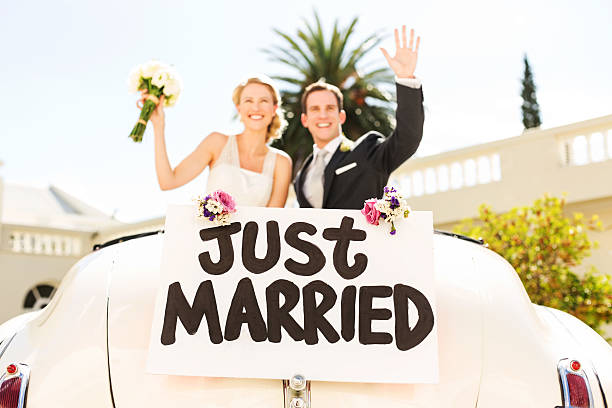 Newlywed couple waving in convertible car with just married sign attached to it. Horizontal shot.