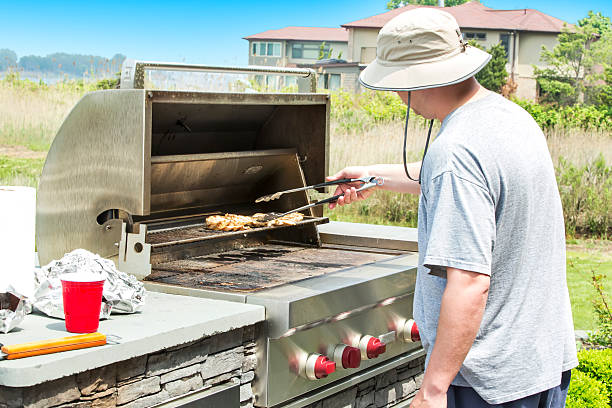Man barbecuing chicken on a outdoor gas grill Caucasian male using tongs to turn chicken breasts that he is barbecuing on a gas grill. His face is not visible. He is wearing a gray t shirt. It is daytime. The chicken is cooking on the upper rack of the grill. Taken with a Canon 5D Mark 3.  rm propane photos stock pictures, royalty-free photos & images