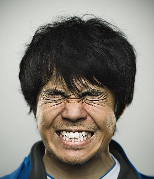 Studio portrait of a japanese young man with closed eyes and clenched teeth. The man has around 30 years and has long black hair and casual clothes. Vertical color image from a medium format digital camera. Sharp focus on eyes.