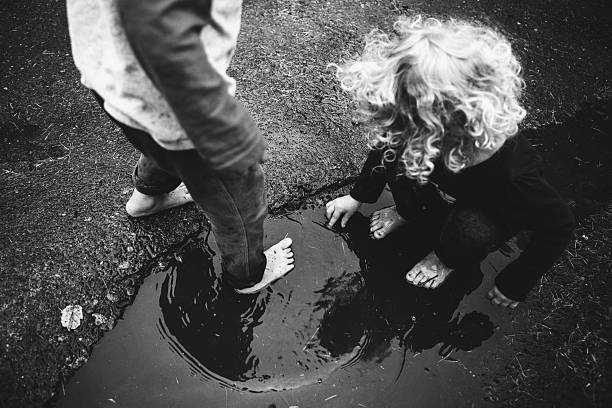 A black and white image of two kids playing as it should be; having fun outdoors getting dirty.  They run and splash in muddy puddles of water in their driveway.  Horizontal image with copy space.