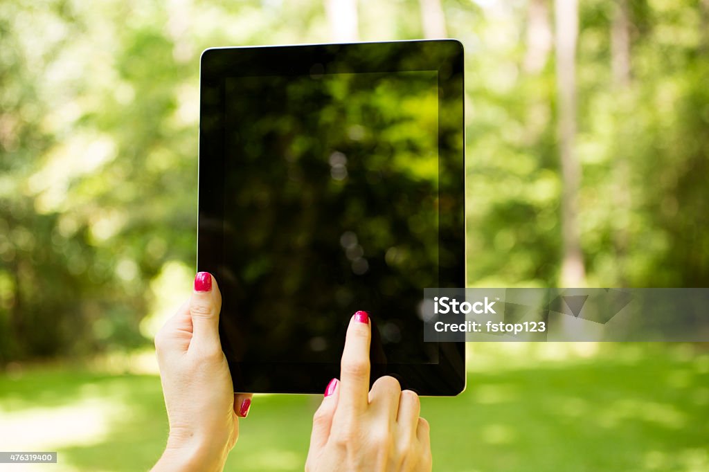 Woman holds digital tablet to take photo of summer landscape. A woman is using her digital tablet to take a photo or video of a summer landscape scene.  She is holding her mobile device to snap a quick photograph of the beautiful green view.  Grassy yard with forest area beyond.  Only her hands are visible. Digital Tablet Stock Photo