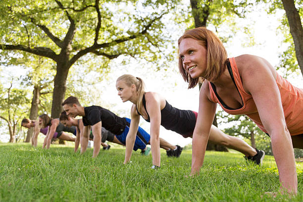 A group of five women and one man exercise together outdoors in a park.  The man is the instructor leading the group in a boot camp style workout.  They are in the grass doing pushups.  Springtime, lots of green in the composition.  Shot in Atlanta, Georgia.