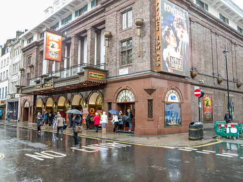 London, UK - May 14, 2015: Tourists standing and walking in the rain in London theatreland district street. People are pictured outside a theatre