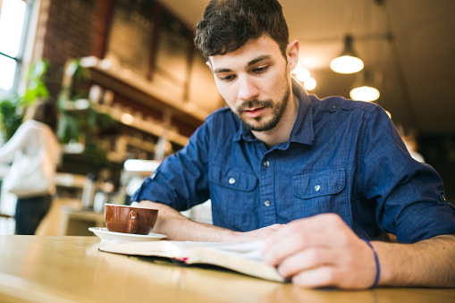 A man concentrates with a serious face as he reads the Bible or a book in his favorite coffee shop, a latte he's drinking sitting next too him on the table.  Horizontal image with copy space.