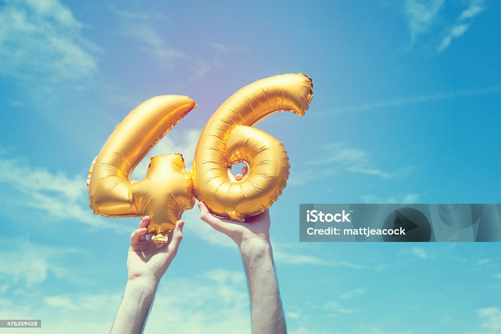 Gold number 46 balloon A gold foil number 46 balloon is held high in the air by caucasian male hand.  The image has been taken outdoors on a bright sunny day, the sky is blue with some clouds. A vintage style effects has been added to the image. 45-49 Years Stock Photo