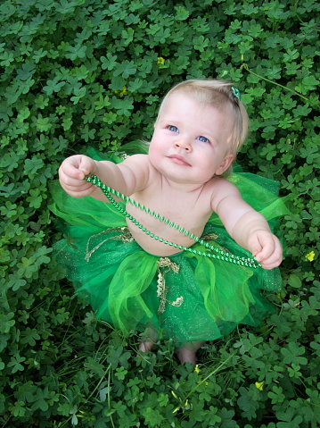 A cute baby girl in a green tutu plays with her green necklace while sitting in a patch of clovers for St. Patrick's Day.