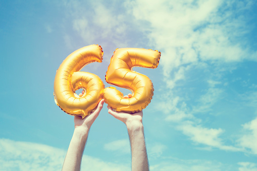 A gold foil number 65 balloon is held high in the air by caucasian male hand.  The image has been taken outdoors on a bright sunny day, the sky is blue with some clouds. A vintage style effects has been added to the image.