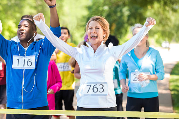 Happy runners celebrating as they win marathon or 5k race African American mid adult man and mature adult Caucasian woman are smiling and celebrating as they cross yellow finish line tape. Runners are winning marathon or 5k race for charity, and are wearing athletic clothing. They are followed by diverse race contestants. All runners are wearing numbers on their shirts. walking for a cause stock pictures, royalty-free photos & images