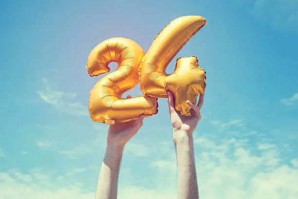 A gold foil number 24 balloon is held high in the air by caucasian male hand.  The image has been taken outdoors on a bright sunny day, the sky is blue with some clouds. A vintage style effects has been added to the image.