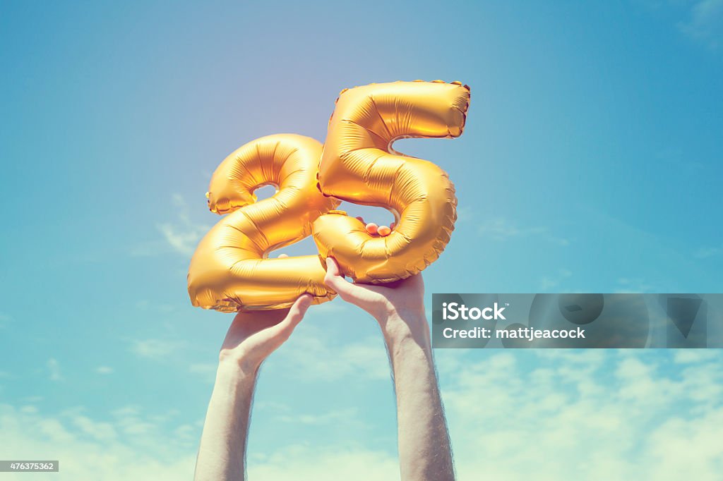 Gold number 25 balloon A gold foil number 25 balloon is held high in the air by caucasian male hand.  The image has been taken outdoors on a bright sunny day, the sky is blue with some clouds. A vintage style effects has been added to the image. 25-29 Years Stock Photo