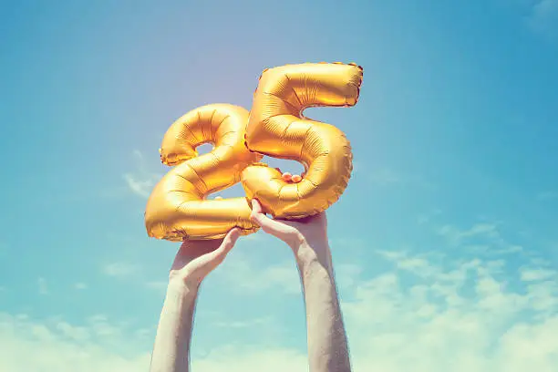 A gold foil number 25 balloon is held high in the air by caucasian male hand.  The image has been taken outdoors on a bright sunny day, the sky is blue with some clouds. A vintage style effects has been added to the image.