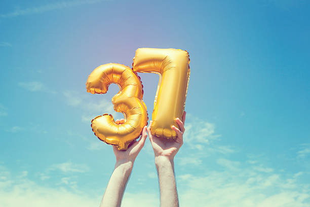 Gold number 37 balloon A gold foil number 37 balloon is held high in the air by caucasian male hand.  The image has been taken outdoors on a bright sunny day, the sky is blue with some clouds. A vintage style effects has been added to the image. number 37 stock pictures, royalty-free photos & images