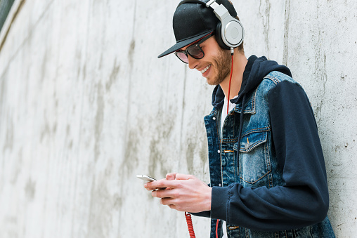 Smiling young man in headphones holding mobile phone while leaning at the concrete wall