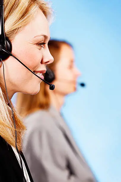 This beautiful young blonde woman wearing a headset smiles happily, as does her colleague behind her. They really enjoy being call-center employees!